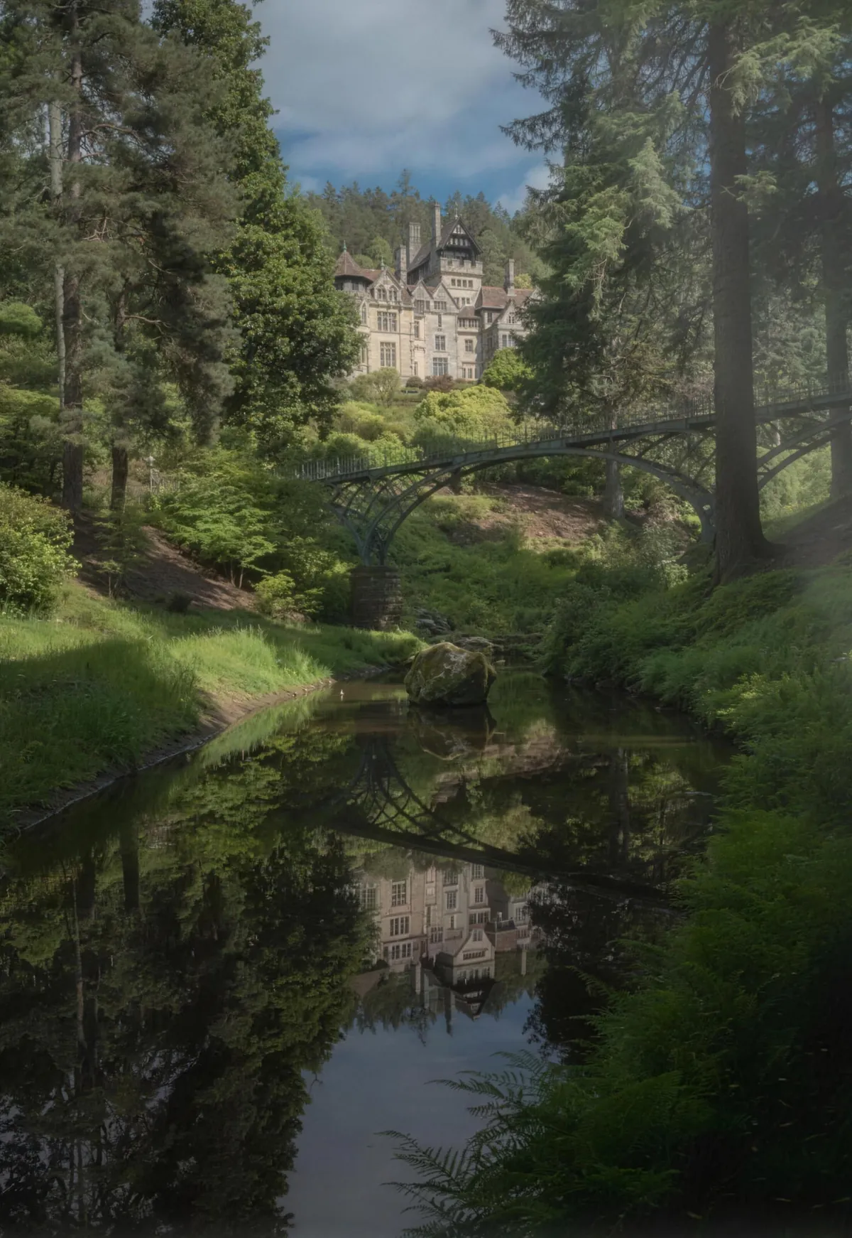 Cragside House above the pines and the iron bridge, reflected in the stream below