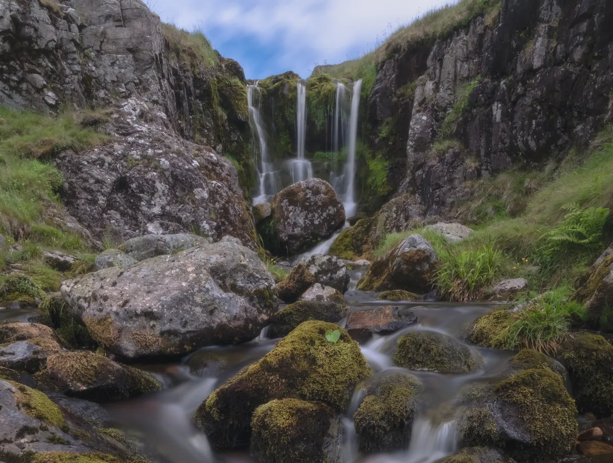 The Three Sisters waterfall falling into the rocky gorge of the Hen Hole, with a stream running over mossy boulders
